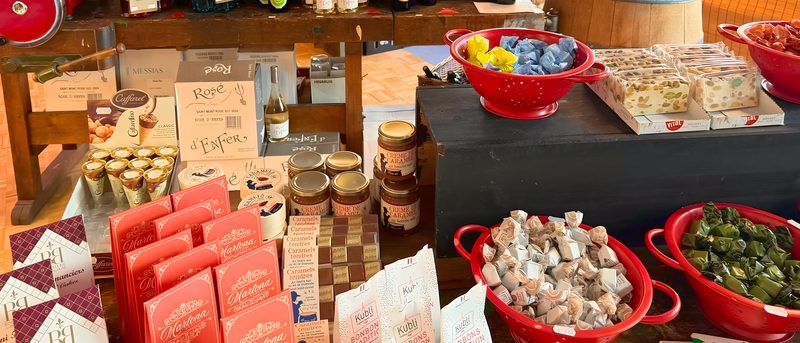 Verkaufstisch mit Pralinen, Gebäck und Feinkost in Gläsern, dahinter Weinflaschen