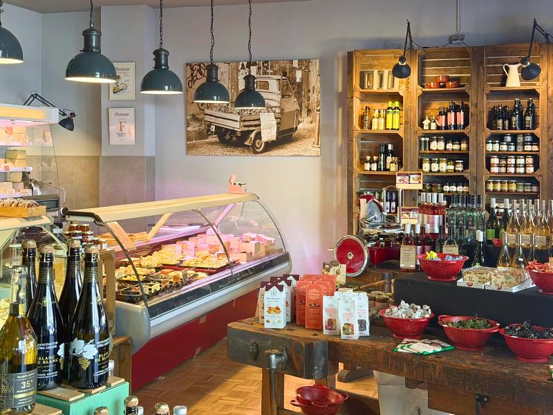 Blick in den Laden: Käsetheke, Hängelampen und Regale mit Feinkost und Wein